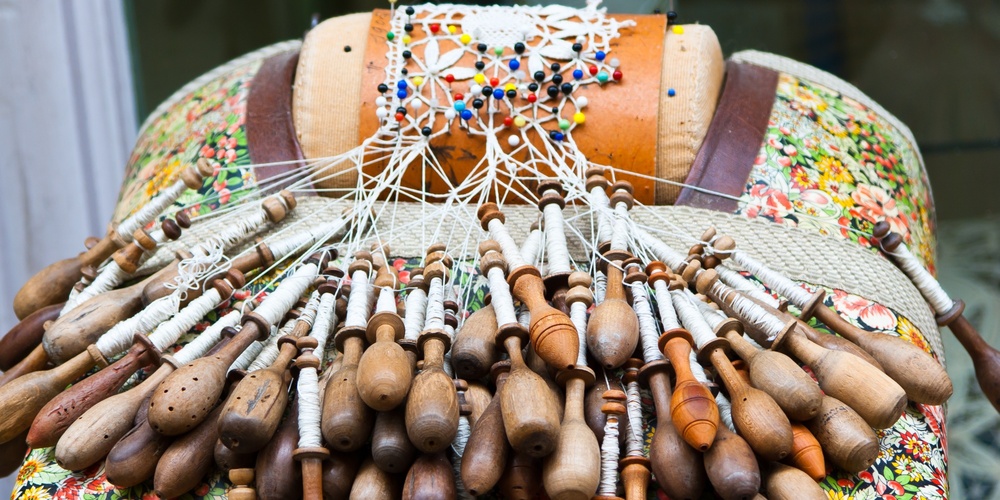 Närbild av en spetskudde med många träspolar och pågående knypplingsarbete.