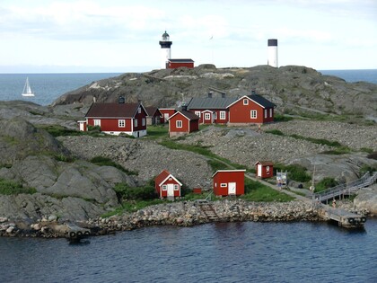 Flera röda stugor och en fyr på en klippig ö vid havet, med en segelbåt i bakgrunden.