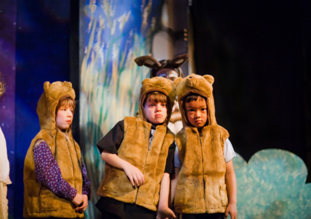 Tre barn i björndräkter står på en scen under en teaterföreställning, med en bakgrund av målade träd.