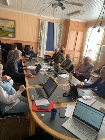 En grupp vuxna sitter runt ett konferensbord med bärbara datorer i ett ljust mötesrum.