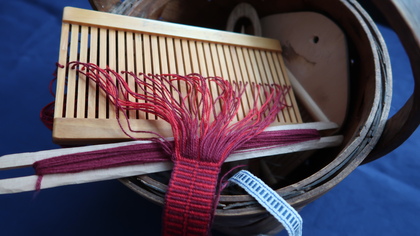 Närbild av ett bandvävningsredskap med rött garn och trädetaljer på blå bakgrund.