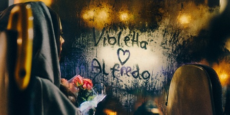 En person med huva sitter på en buss, håller blommor och tittar på ett immigt fönster där det står 'Violetta ♥ Alfredo'.