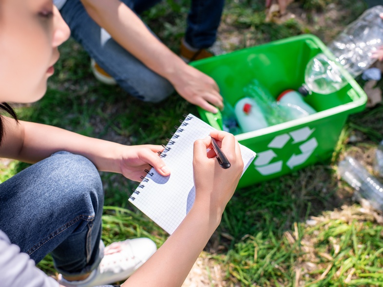 Person skriver i anteckningsblock bredvid återvinningslåda med plastflaskor utomhus.