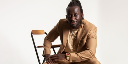 En person i beige kostym sitter på en stol mot en vit bakgrund.