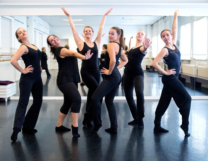 Sex kvinnor i svarta kläder poserar i en dansstudio framför en spegel.