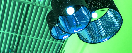 Flera taklampor med industriell design hänger från ett tak med synliga balkar, belysta i grönt och blått ljus.