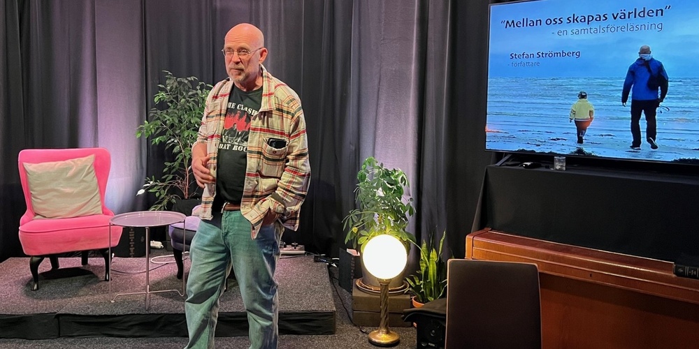 Man står på en scen med svart bakgrund, bredvid en TV-skärm med text och en bild på två personer vid havet.