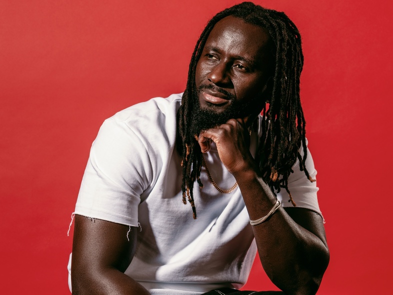 Pa Modou Badjie i vit t-shirt och svarta arbetsbyxor sitter på högtalare mot röd bakgrund