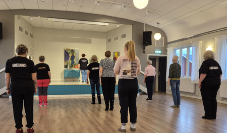 En grupp personer står i ett dansrum och följer en instruktör på en liten scen längst fram.