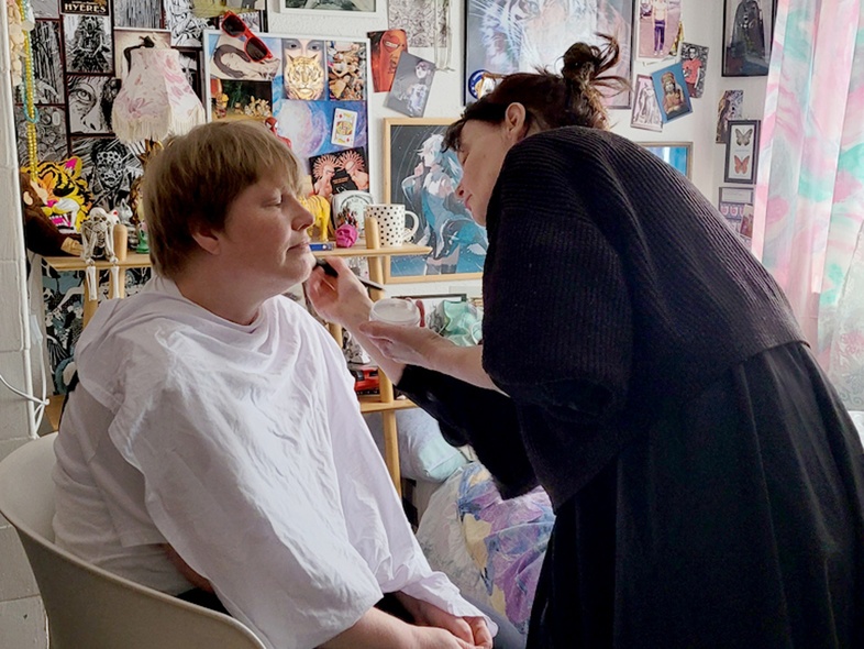 En kvinna sitter i en stol och blir sminkad inför en filminspelning. 