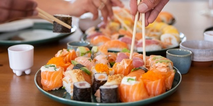 Två personer plockar sushi med ätpinnar från ett fat på ett matbord.