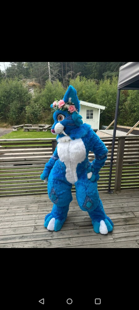 Person i blått fursuit-kostym med blommor på huvudet står på en altan utomhus.