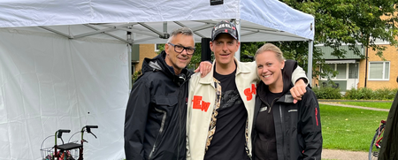 Tre personer står tillsammans och ler framför ett vitt tält på en gräsmatta. En rullator syns i bakgrunden.