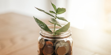 En glasburk fylld med mynt står på ett träbord, med en grön växt som växer ur burken.