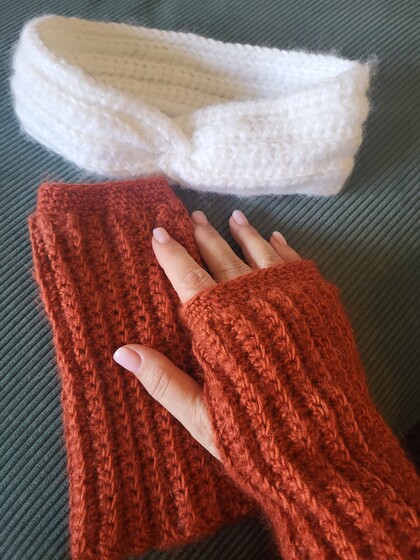 En hand med fingerlösa stickade vantar i rostfärg, bredvid ett vitt stickat pannband på en grön tygsoffa.