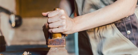 En person i arbetskläder hyvlar trä i en snickarverkstad, fokus på händer och verktyg.