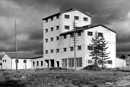 En svartvit bild på ett stort vitt hus med fyra våningar. En mörkt molning himmel i bakgrunden. Framför huset telefonstolpar med kablar. 