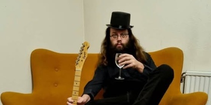 Person med långt hår och hatt sitter på en orange soffa med en elgitarr och ett vinglas.