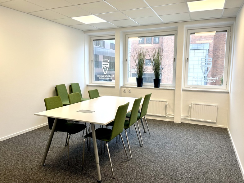 Ljust mötesrum med vitt bord, sex gröna stolar och två fönster med växter i fönsterkarmen.
