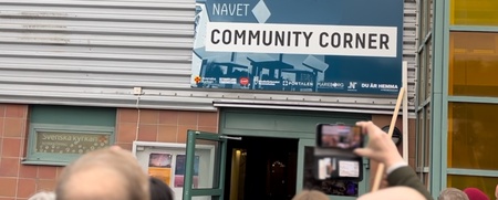 En grupp människor står utanför en byggnad med skylten 'Community Corner' ovanför ingången.