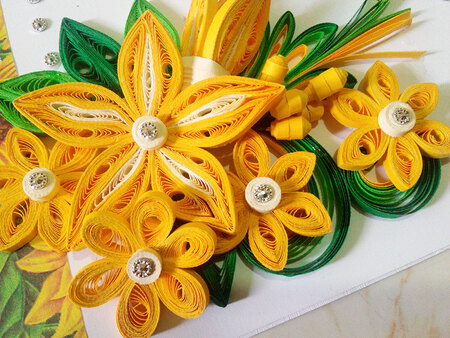 Färgglada blommor skapade av quillingpapper i gult, vitt och grönt på ett vitt underlag.
