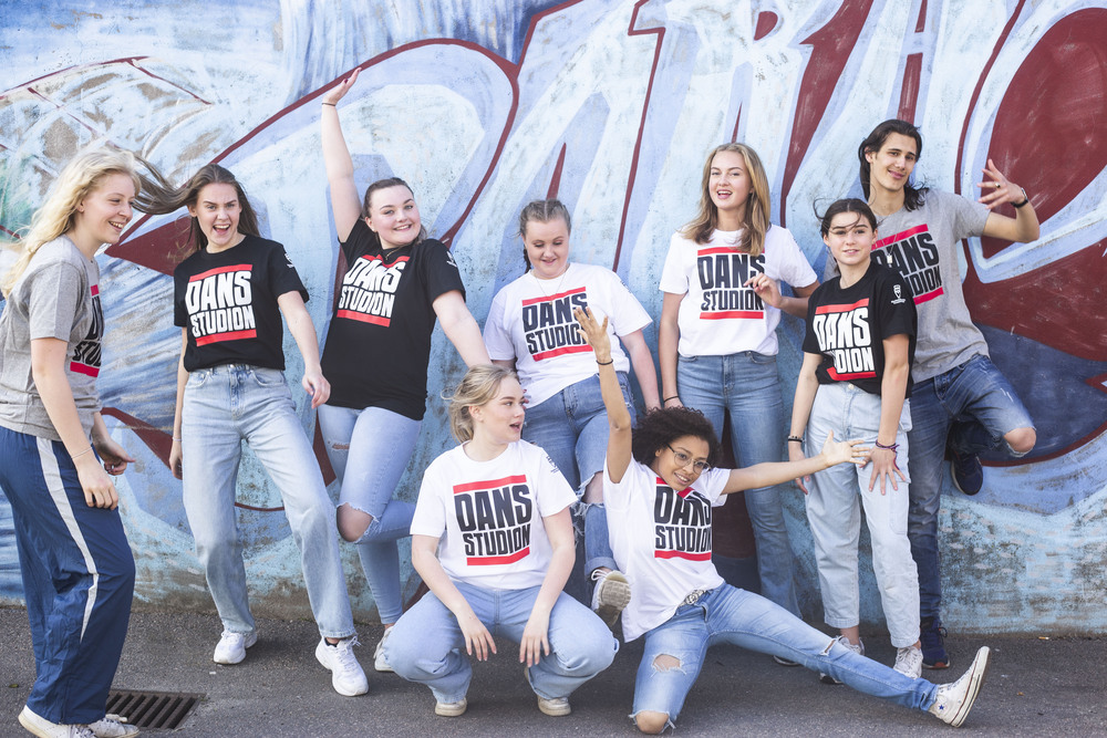 En grupp unga personer poserar glatt framför en graffitivägg, alla bär t-shirts med texten 'DANS STUDION'.