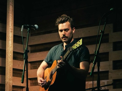 Man spelar akustisk gitarr på en scen med träväggar och mikrofoner.