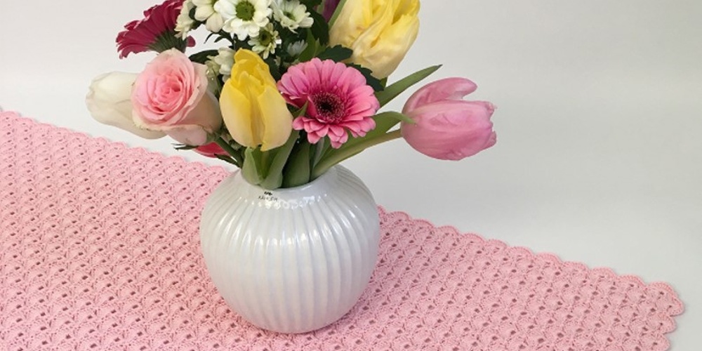 Vit vas med färgglada blommor står på en rosa virkad duk mot vit bakgrund.