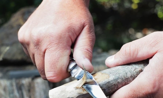 Två händer täljer en träpinne med en fällkniv utomhus.