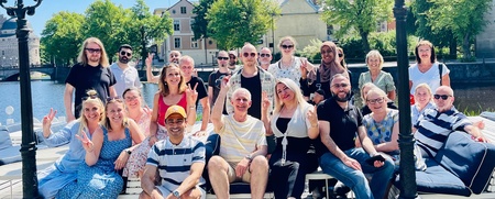 En grupp människor sitter och står på en brygga vid vattnet, omgivna av lampstolpar och träd, en solig dag.