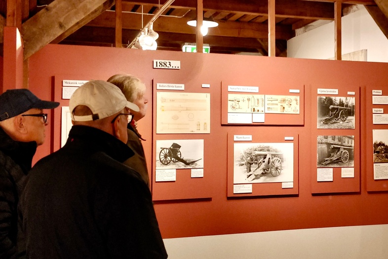 Fem personer tittar på historiska utställningstavlor i ett museum med träbjälkar och rödbruna väggar.