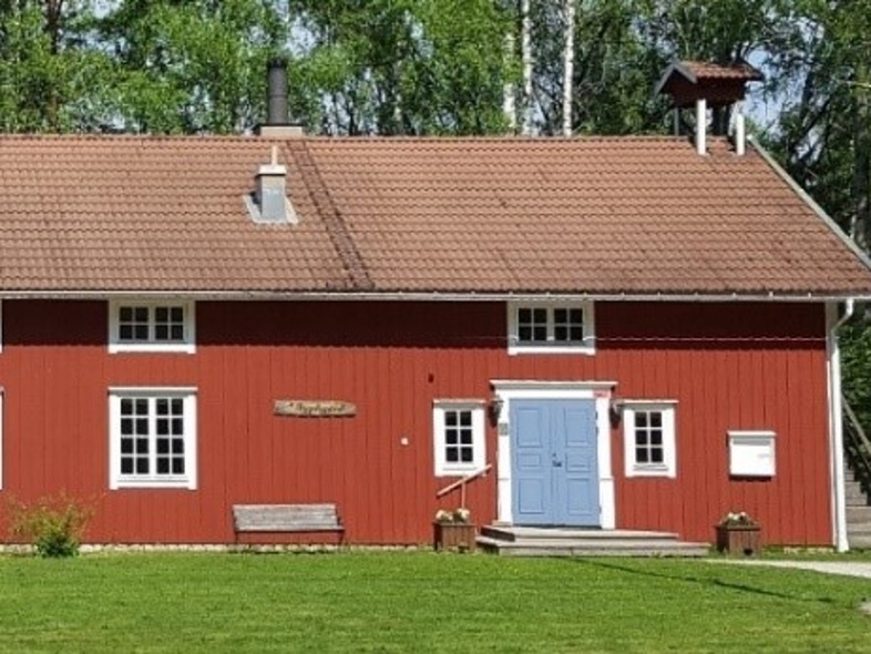 Rött trähus med vita knutar och blå dörrar, omgiven av gräsmatta och träd.