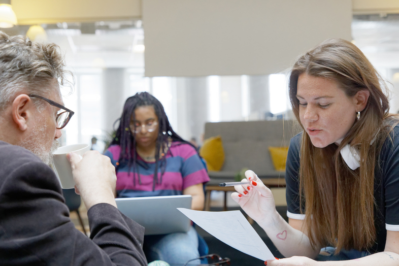 Tre personer arbetar tillsammans i ett modernt kontorsutrymme, två diskuterar papper, en använder laptop i bakgrunden.