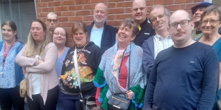 En grupp människor står och sitter utomhus framför en tegelvägg, några med färgglada kläder.