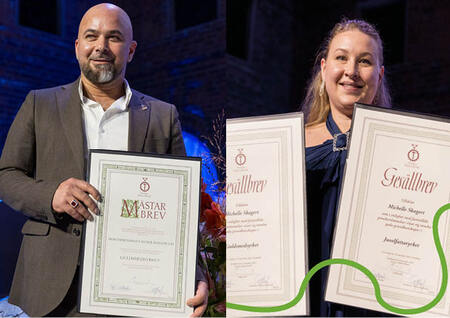 Två personer håller diplom och poserar leende på en scen med mörk bakgrund.