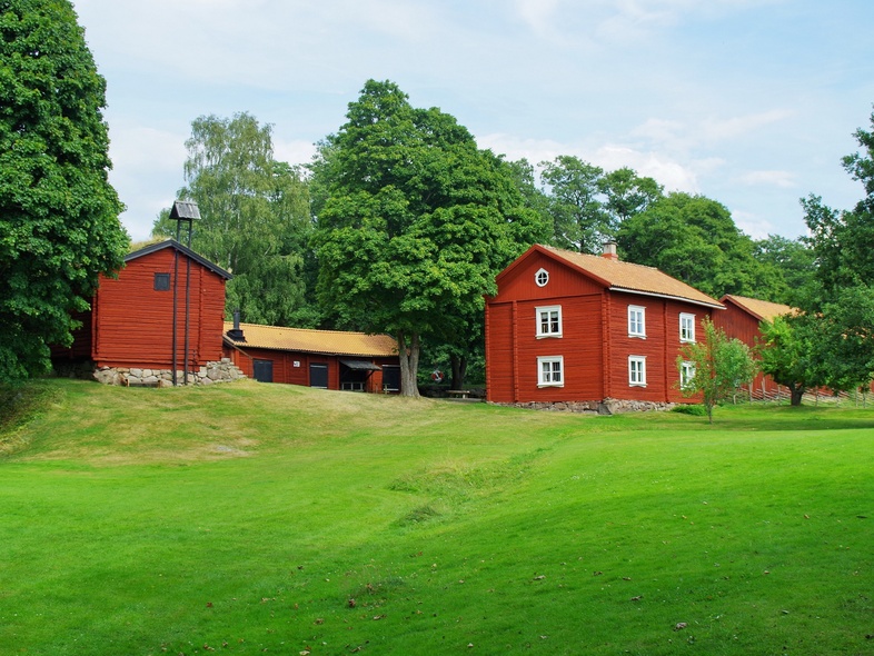 Röda trähus omgivna av grönskande träd och gräsmatta under blå himmel.