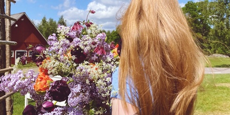 En person med långt blont hår håller en stor bukett blommor utomhus, ryggen vänd mot kameran.