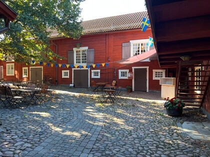 En innergård med kullersten, röda trähus, utemöbler och svenska flaggor i sommarsol.