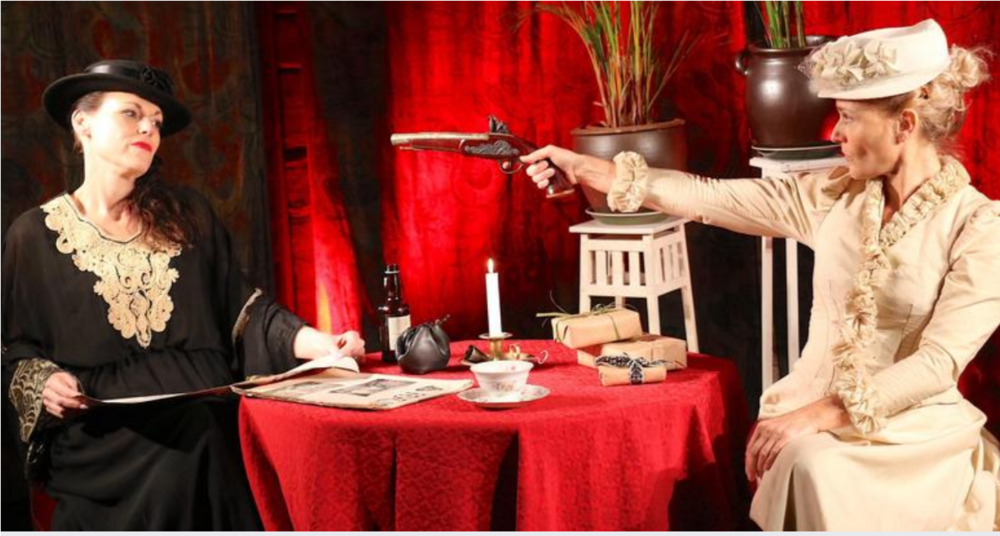 Två kvinnor i vintagekläder sitter vid ett bord, en riktar en pistol mot den andra.