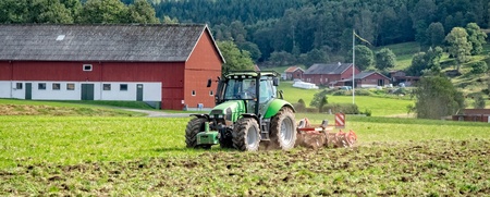 En grön traktor plöjer en åker framför en röd ladugård på landsbygden med skog i bakgrunden.