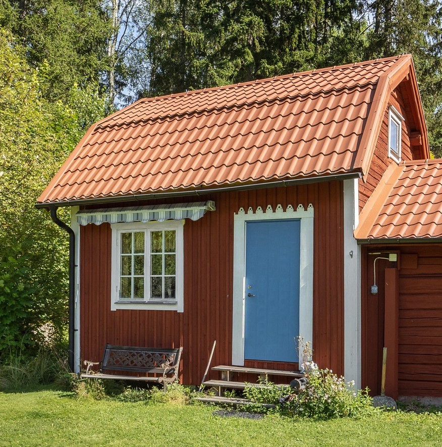 Liten röd stuga med blå dörr, vitt fönster och tegeltak i grönskande trädgård.