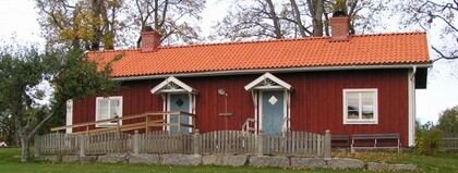 Rött trähus med vita knutar, två blå dörrar, staket och tegeltak, omgiven av träd och gräsmatta.