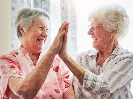 Två äldre kvinnor skrattar och ger varandra en high five i ett ljust rum.