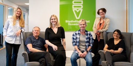 Sex personer poserar leende i ett kontorsutrymme framför en grön skylt med texten 'Studieförbundet Vuxenskolan'.