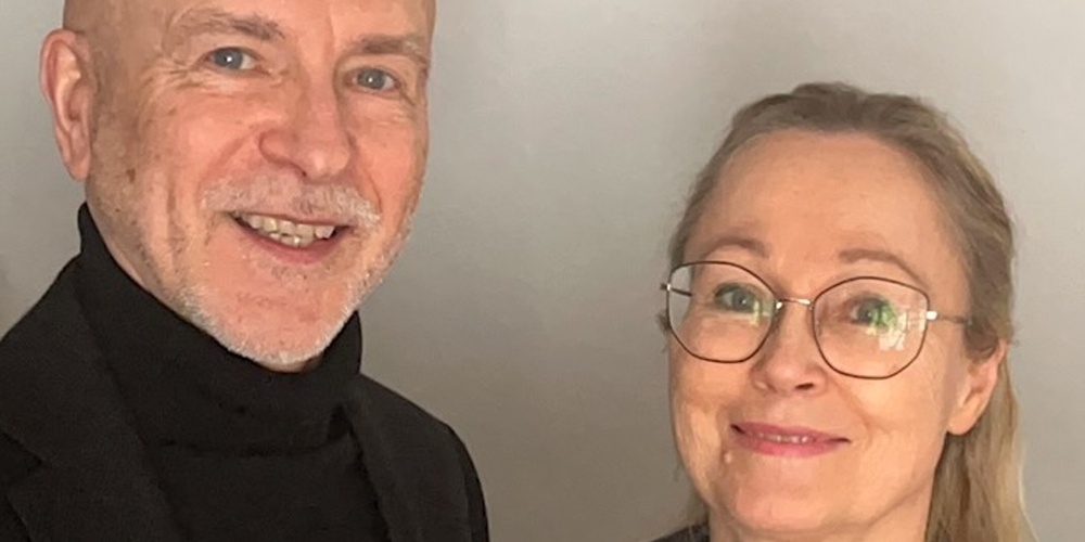 Två personer står bredvid varandra mot en ljus bakgrund, en bär svart kavaj och den andra en svart jacka och grön halskedja.