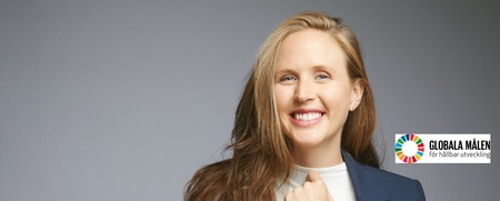 Leende kvinna med långt hår i kavaj och ljus topp, står mot grå bakgrund med logotyp för Globala målen.