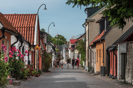 En pittoresk kullerstensgata med färgglada hus, blommor och människor som promenerar en solig dag.
