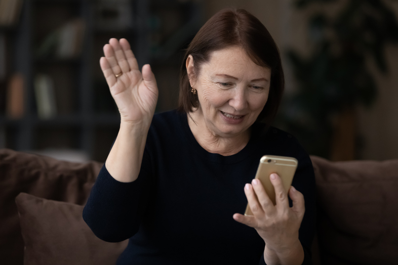 Kvinna med brunt hår vinkar och ler mot en mobiltelefon, sitter i soffa inomhus.