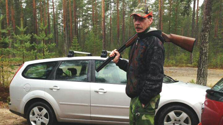 Man med gevär på axeln står bredvid en silverfärgad bil i en skogsmiljö.