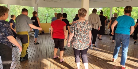 En grupp vuxna dansar tillsammans på ett trägolv i en ljus paviljong med stora fönster.